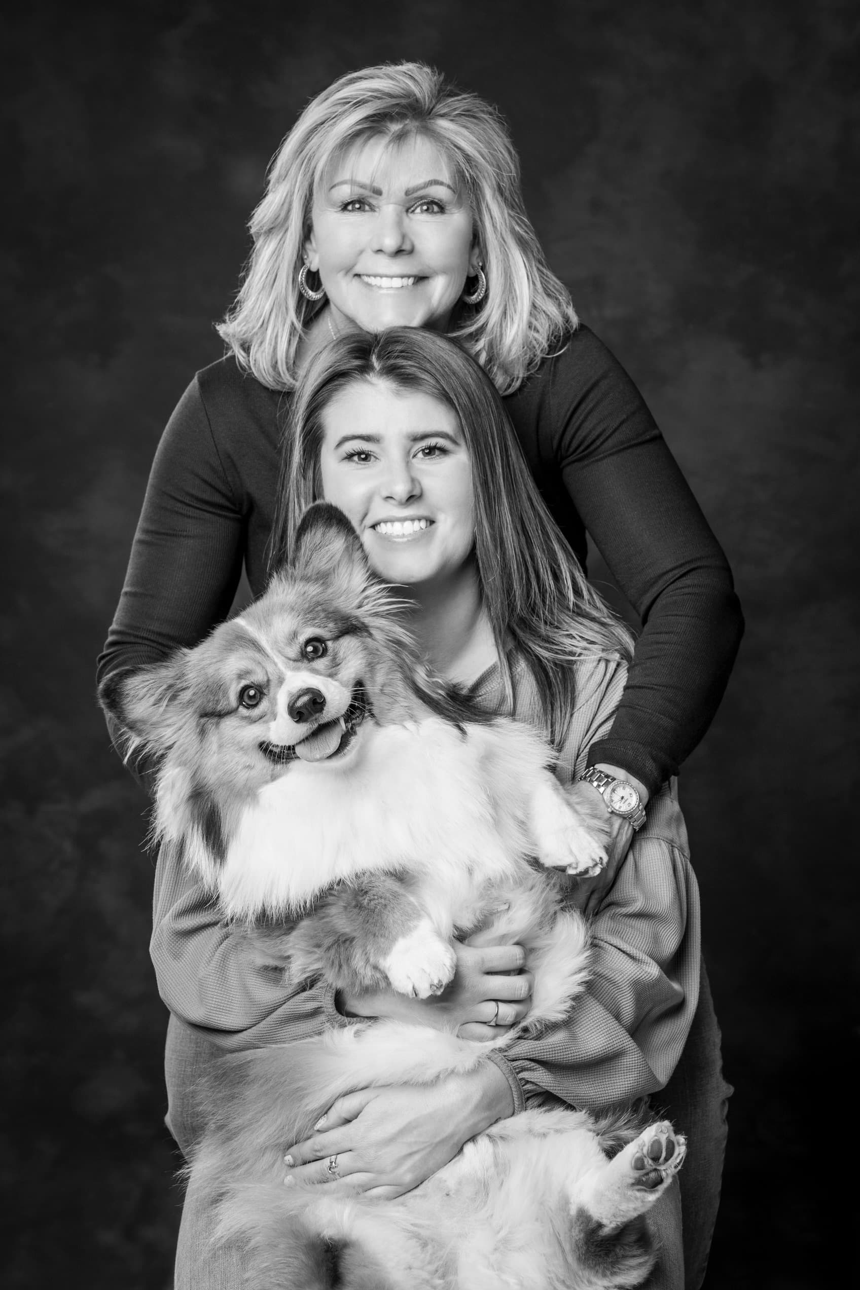 Black-and-white studio portrait of Mary Leeds standing behind her daughter, who is holding a smiling fluffy corgi